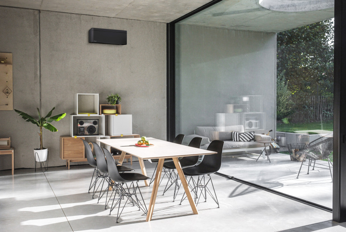 Ein zeitgemäßer Essbereich mit einem eleganten Holztisch, umgeben von schwarzen Stühlen. Die Einrichtung ist minimalistisch gehalten, mit Zimmerpflanzen und einer an der Wand montierten Klimaanlage. Große Glasfenster bieten den Blick auf eine Terrasse mit Sitzgelegenheiten und viel Grün.
