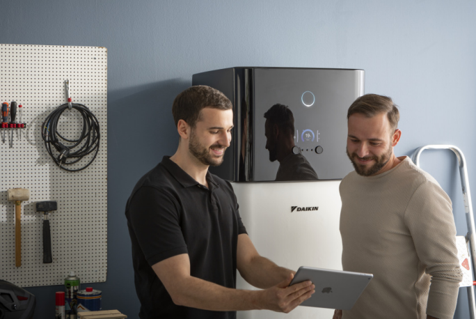 Zwei Personen sind in einer Werkstattumgebung zu sehen und beschäftigen sich mit einem Daikin-HVAC-Gerät. Eine Person hält ein Tablet und prüft möglicherweise Systemeinstellungen oder Diagnosedaten. Im Hintergrund befinden sich Werkzeuge und Ausrüstung, was auf ein professionelles Umfeld hinweist. Das Daikin-Logo ist deutlich auf dem Gerät zu erkennen.